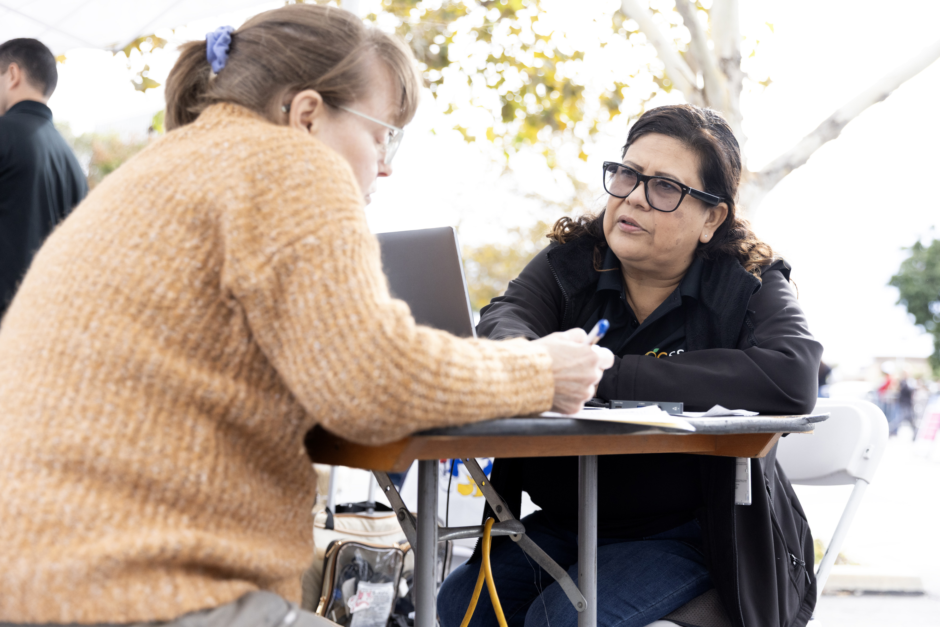 A photo of a Hispanic woman with a laptop at a table outside. A white woman sits at a chair in front of her, writing on a piece of paper.